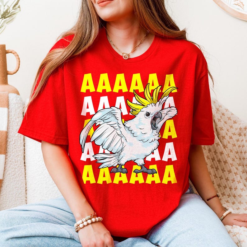 Vibrant red T-shirt featuring a cartoon sulfur-crested cockatoo screaming with its wings spread against a background of repeating "AAAAAA" text, ideal for bird owners, parrot lovers, and fans of chaotic, loud-humor graphic apparel.