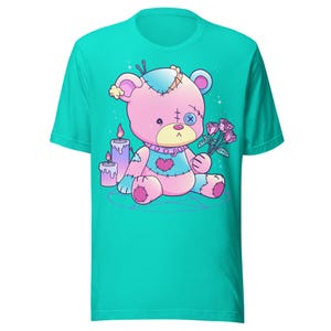 May include: A turquoise t-shirt with a pink and white stitched teddy bear holding a bouquet of pink roses. The teddy bear is sitting on a purple pentagram with two purple candles.