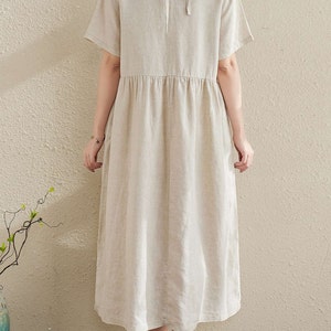 Puede incluir: Un vestido de lino beige con cuello redondo y mangas cortas. El vestido tiene un lazo en la parte posterior del cuello y una cintura fruncida.