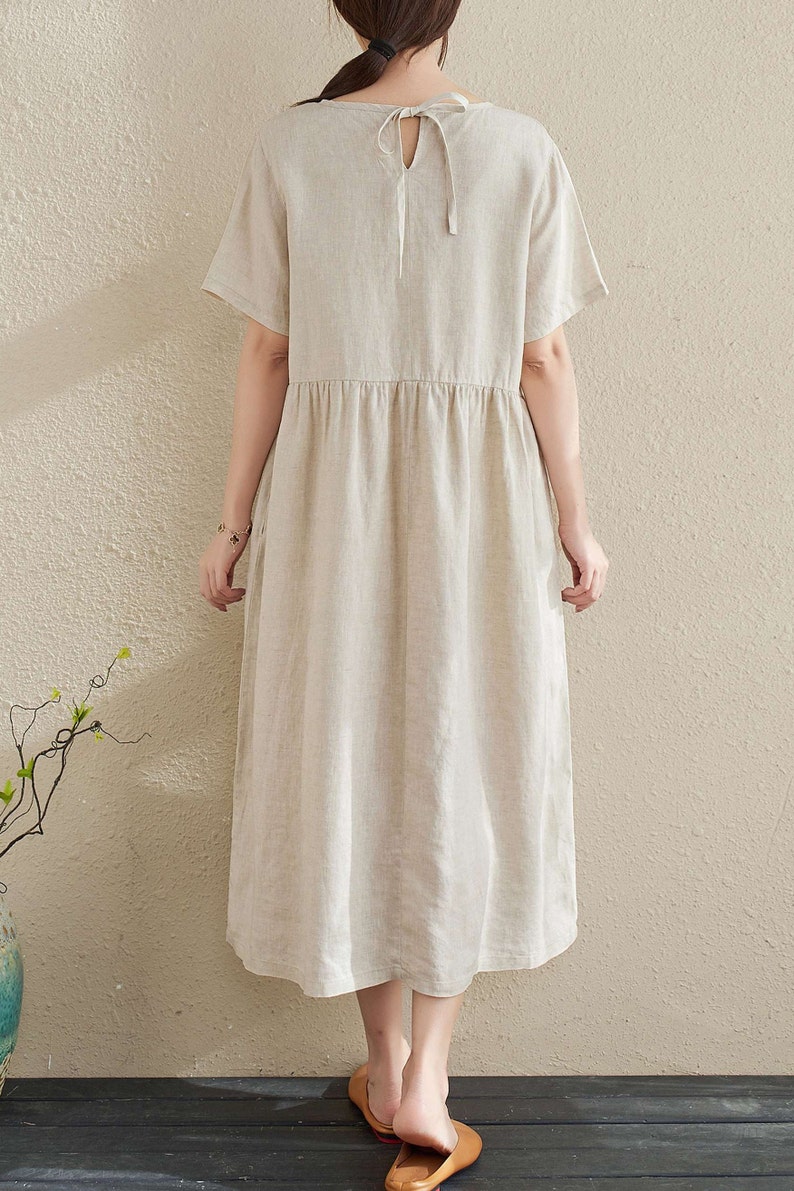 Puede incluir: Un vestido de lino beige con cuello redondo y mangas cortas. El vestido tiene un lazo en la parte posterior del cuello y una cintura fruncida.