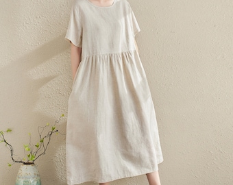 May include: A simple, loose-fitting, beige linen dress with short sleeves and a round neckline. The dress has a gathered waist and falls to the mid-calf.