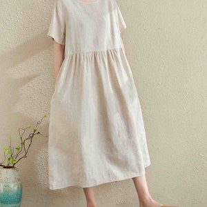 Puede incluir: Un vestido de lino beige con mangas cortas y cintura fruncida. El vestido tiene un corte holgado y llega a la mitad de la pantorrilla.