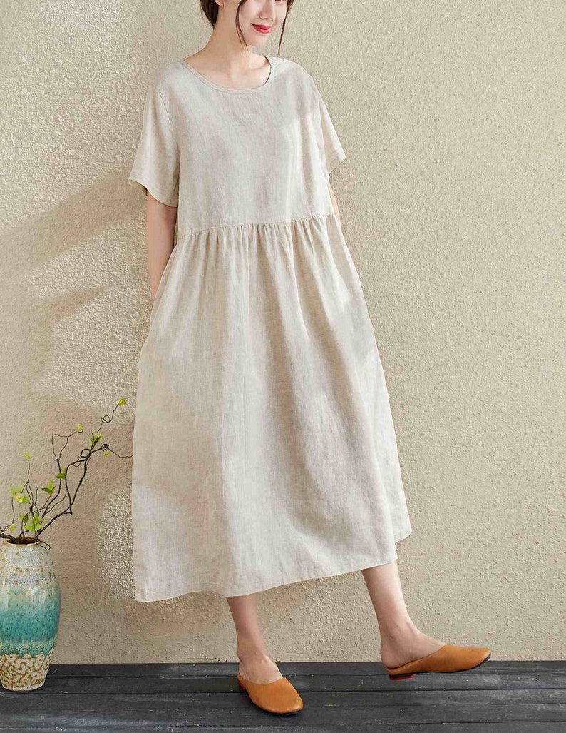 Puede incluir: Un vestido de lino beige con mangas cortas y cintura fruncida. El vestido tiene un corte holgado y llega a la mitad de la pantorrilla.