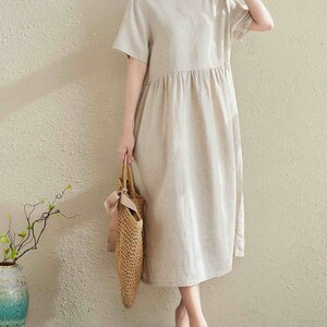 Puede incluir: Un vestido de lino beige con cuello redondo y mangas cortas. El vestido tiene una cintura fruncida y llega a la mitad de la pantorrilla. La modelo lleva el vestido con sandalias de cuero marr&oacute;n y un bolso de paja.