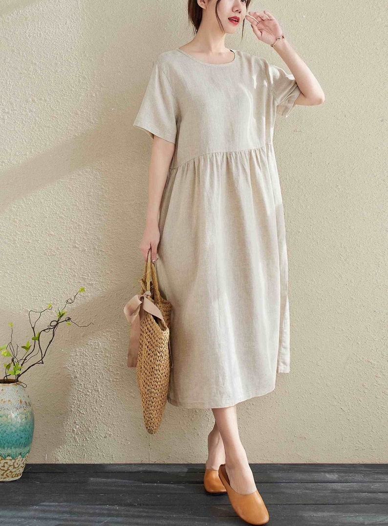 Puede incluir: Un vestido de lino beige con cuello redondo y mangas cortas. El vestido tiene una cintura fruncida y llega a la mitad de la pantorrilla. La modelo lleva el vestido con sandalias de cuero marr&oacute;n y un bolso de paja.