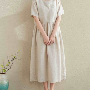 Puede incluir: Un vestido de lino beige con cuello redondo y mangas cortas. El vestido tiene una cintura fruncida y llega a la mitad de la pantorrilla.