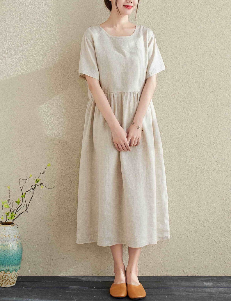 Puede incluir: Un vestido de lino beige con cuello redondo y mangas cortas. El vestido tiene una cintura fruncida y llega a la mitad de la pantorrilla.
