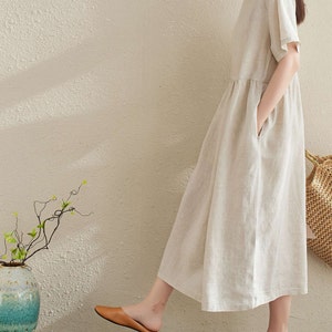 Puede incluir: Un vestido de lino beige de manga corta y corte holgado. El vestido tiene bolsillos a los lados y una cintura fruncida. La modelo lleva el vestido con sandalias marrones.