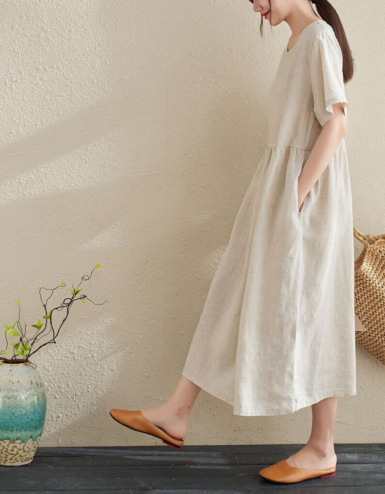 Puede incluir: Un vestido de lino beige de manga corta y corte holgado. El vestido tiene bolsillos a los lados y una cintura fruncida. La modelo lleva el vestido con sandalias marrones.