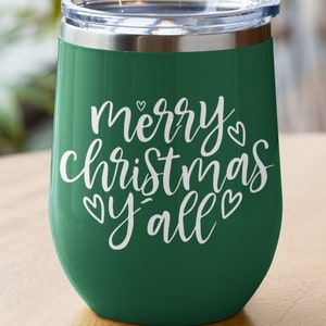 May include: A green insulated wine tumbler with a clear lid. The tumbler has the words "merry christmas y'all" in white script. The tumbler is on a wooden surface.