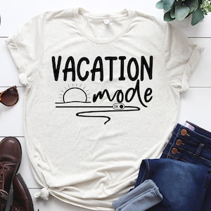 以下が含まれることがあります： 白いTシャツに黒い文字で「Vacation Mode」と書かれています。テキストには、水面に沈む夕日と、「ON」と書かれたスイッチのグラフィックが添えられています。