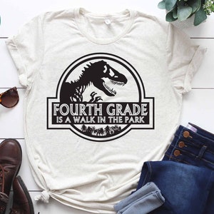 May include: A white t-shirt with a black graphic of a dinosaur silhouette inside a circle. The text "FOURTH GRADE IS A WALK IN THE PARK" is below the dinosaur.