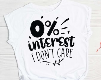 Puede incluir: Camiseta blanca con texto negro que dice "0% de interés no me importa". La camiseta está anudada en la parte delantera y tiene un pequeño diseño floral en la parte inferior. Una decoración en forma de corazón rojo está sobre una superficie de madera blanca.