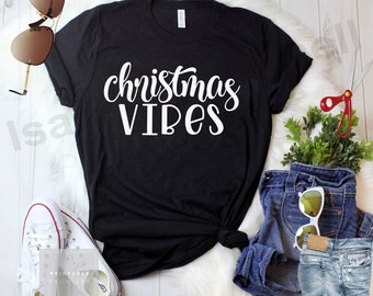 Könnte beinhalten: Schwarzes T-Shirt mit dem weißen Schriftzug "Christmas VIBES". Das Shirt ist mit blauen Jeans, weißen Turnschuhen und Sonnenbrillen gestylt. Eine Schere und eine kleine Pflanze sind ebenfalls zu sehen.