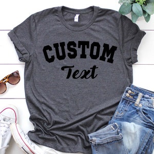 May include: A dark gray t-shirt with the text "CUSTOM Text" in black lettering. The shirt is laid flat on a white wooden surface with a pair of sunglasses and a pair of white sneakers.