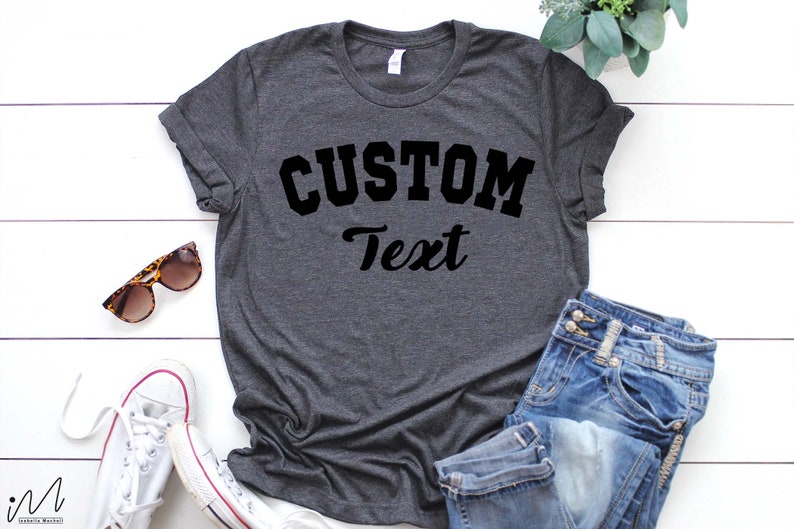May include: A dark gray t-shirt with the text "CUSTOM Text" in black lettering. The shirt is laid flat on a white wooden surface with a pair of sunglasses and a pair of white sneakers.