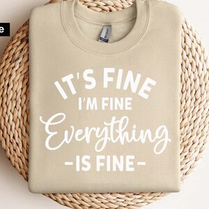 May include: A sand-colored crewneck sweatshirt featuring the white printed text "IT'S FINE I'M FINE Everything IS FINE" centered on the chest. The garment has long sleeves, a ribbed collar, and a standard fit, presented as a digital file design.
