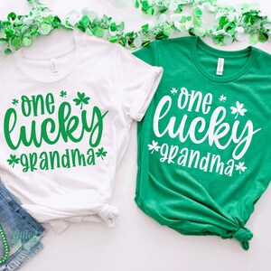 K&ouml;nnte beinhalten: Zwei wei&szlig;e T-Shirts mit gr&uuml;nen Kleeblatt-Designs. Der Text auf den Shirts lautet "one lucky grandma".