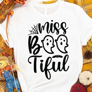 May include: White t-shirt with a black graphic that says "miss Boo-tiful" with two ghost figures and a spiderweb. The shirt is displayed on a yellow plaid blanket with fall-themed decorations.