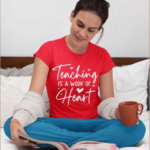 May include: A woman wearing a red t-shirt with the text "Teaching is a work of heart" in white lettering. She is sitting on a bed with a magazine in her lap and a cup of coffee in her hand.