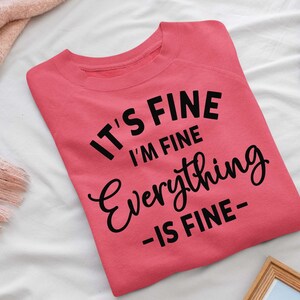 May include: A coral-colored crewneck sweatshirt featuring black text that reads IT'S FINE I'M FINE Everything -IS FINE- in a mix of bold sans-serif and cursive fonts. The garment is folded and displayed flat, showcasing the front graphic design.