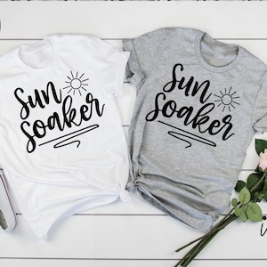 Peut inclure: Deux t-shirts blancs avec le texte "Sun Soaker" en &eacute;criture noire avec un petit graphique de soleil. Les chemises sont dispos&eacute;es sur une surface en bois blanc avec un bouquet de fleurs roses et blanches.