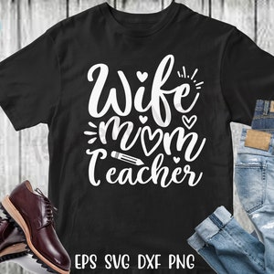 Pu&ograve; includere: Maglietta nera con testo bianco che recita "Wife Mom Teacher" con cuori e una grafica a matita. La maglietta &egrave; su uno sfondo di legno con jeans.