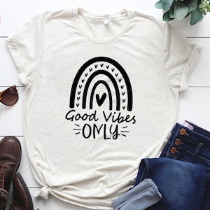 Peut inclure: Un t-shirt blanc avec un motif arc-en-ciel noir et le texte "Good Vibes Only".