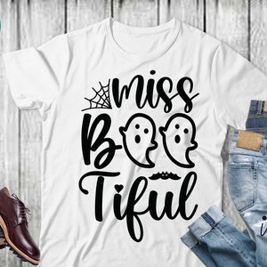 May include: White t-shirt with the phrase "miss Boo-tiful" in black script and two ghost illustrations. The shirt is paired with blue jeans and brown leather shoes, suggesting a Halloween theme.