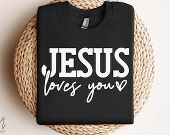 Può includere: Felpa nera con la scritta bianca "JESUS loves you". La felpa è appoggiata su una superficie circolare intrecciata. Il testo è in grassetto, a blocchi, con un "loves you" in corsivo e un simbolo a cuore.