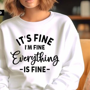 May include: A white crewneck sweatshirt featuring black text that reads IT'S FINE I'M FINE Everything -IS FINE- printed on the front. The garment has long sleeves and a ribbed neckline. The text is displayed in a mix of bold block and script fonts.
