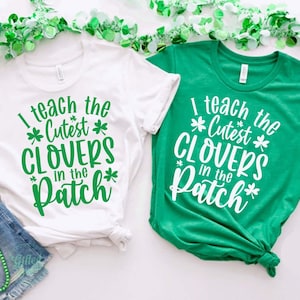 May include: Two green and white t-shirts with the text "I teach the cutest clovers in the patch" on them. The shirts are on a white background with green garland.