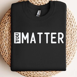 May include: Black crew neck sweatshirt with the white text "YOU MATTER". The sweatshirt is displayed on a woven, circular, beige surface. The Gildan brand tag is visible on the collar.