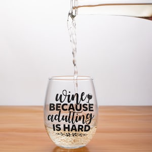 May include: A clear glass wine glass with the text "wine because adulting is hard" printed on it. Wine is being poured into the glass from a bottle.