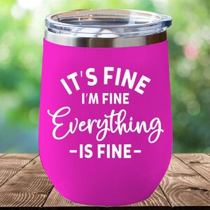 May include: A bright pink stainless steel tumbler with a clear lid, featuring the white printed text IT'S FINE I'M FINE Everything IS FINE. The tumbler has a rounded shape and a metallic silver rim at the top, designed for holding beverages.