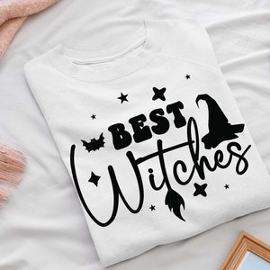 Puede incluir: Sudadera blanca con texto negro que dice "Best Witches" con un sombrero de bruja y un gráfico de escoba.