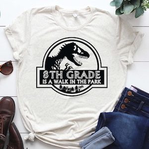 May include: A white t-shirt with a black graphic of a dinosaur and the text "8th Grade is a walk in the park".