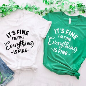 May include: Two short-sleeved t-shirts, one white with black text and one green with white text. Both shirts feature the printed phrase IT'S FINE I'M FINE Everything IS FINE. The shirts are displayed alongside green beads and denim shorts.