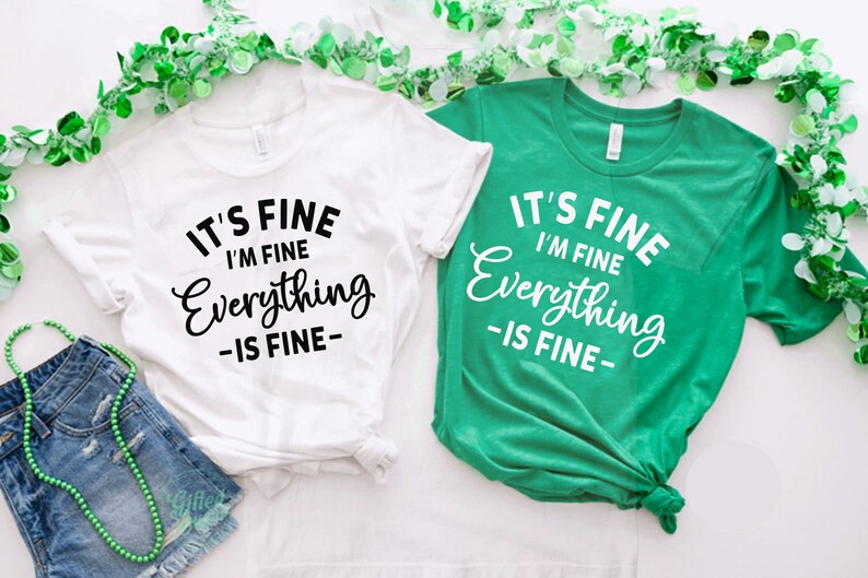 May include: Two short-sleeved t-shirts, one white with black text and one green with white text. Both shirts feature the printed phrase IT'S FINE I'M FINE Everything IS FINE. The shirts are displayed alongside green beads and denim shorts.