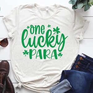 May include: A white t-shirt with a green design that says "*one* lucky *para*" with shamrock symbols around the text. The shirt is laid out on a white wooden surface with a pair of jeans and brown boots.