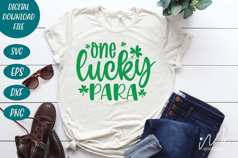May include: A white t-shirt with a green design that says "*one* lucky *para*" with shamrock symbols around the text. The shirt is laid out on a white wooden surface with a pair of jeans and brown boots.