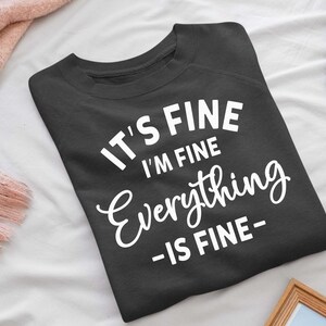 May include: A dark charcoal gray crewneck sweatshirt featuring white screen-printed text that reads IT'S FINE I'M FINE Everything -IS FINE- in a combination of bold sans-serif and cursive script fonts centered on the front of the garment.