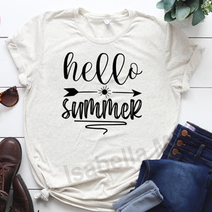 Peut inclure: Un t-shirt blanc avec le texte "hello summer" en lettres cursives noires. Une fl&egrave;che noire avec un graphique de soleil pointe vers la droite. Le t-shirt est pos&eacute; sur une surface en bois blanc avec une paire de lunettes de soleil, des bottes brunes et un jean bleu.