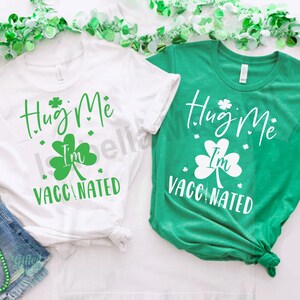 以下が含まれることがあります： 緑のクローバーのデザインと「Hug Me I'm Vaccinated」というテキストが書かれた白いTシャツ2枚。1枚は白、もう1枚は緑です。