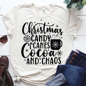 May include: A white t-shirt with black text that reads "Christmas Candy Canes Cocoa and Chaos". The text is surrounded by small stars and a candy cane, a peppermint swirl, and a cup of hot cocoa with whipped cream.
