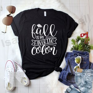 May include: A black t-shirt with white text that reads "fall is my favorite color" with a small leaf graphic above the word "fall". The shirt is laid out on a white surface with a pair of sunglasses, a pair of jeans, and a small potted plant.