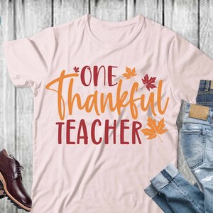 May include: A pink t-shirt with the text "One Thankful Teacher" in orange and red lettering. There are small orange and red leaf designs around the text. The t-shirt is on a wooden surface with a pair of jeans and brown shoes.