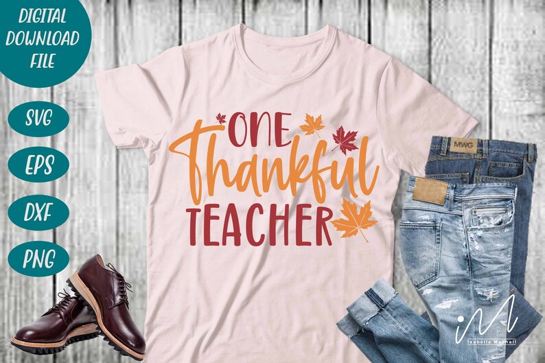 May include: A pink t-shirt with the text "One Thankful Teacher" in orange and red lettering. There are small orange and red leaf designs around the text. The t-shirt is on a wooden surface with a pair of jeans and brown shoes.