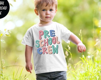 Może przedstawiać: Biała koszulka z napisem "PRE SCHOOL CREW" w kolorowych, bąbelkowych literach. Koszulkę nosi małe dziecko stojące na trawiastym polu.