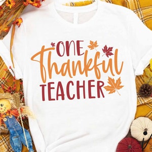 May include: White t-shirt with the text "One Thankful Teacher" in orange and red lettering. The text is surrounded by small red and orange maple leaves.